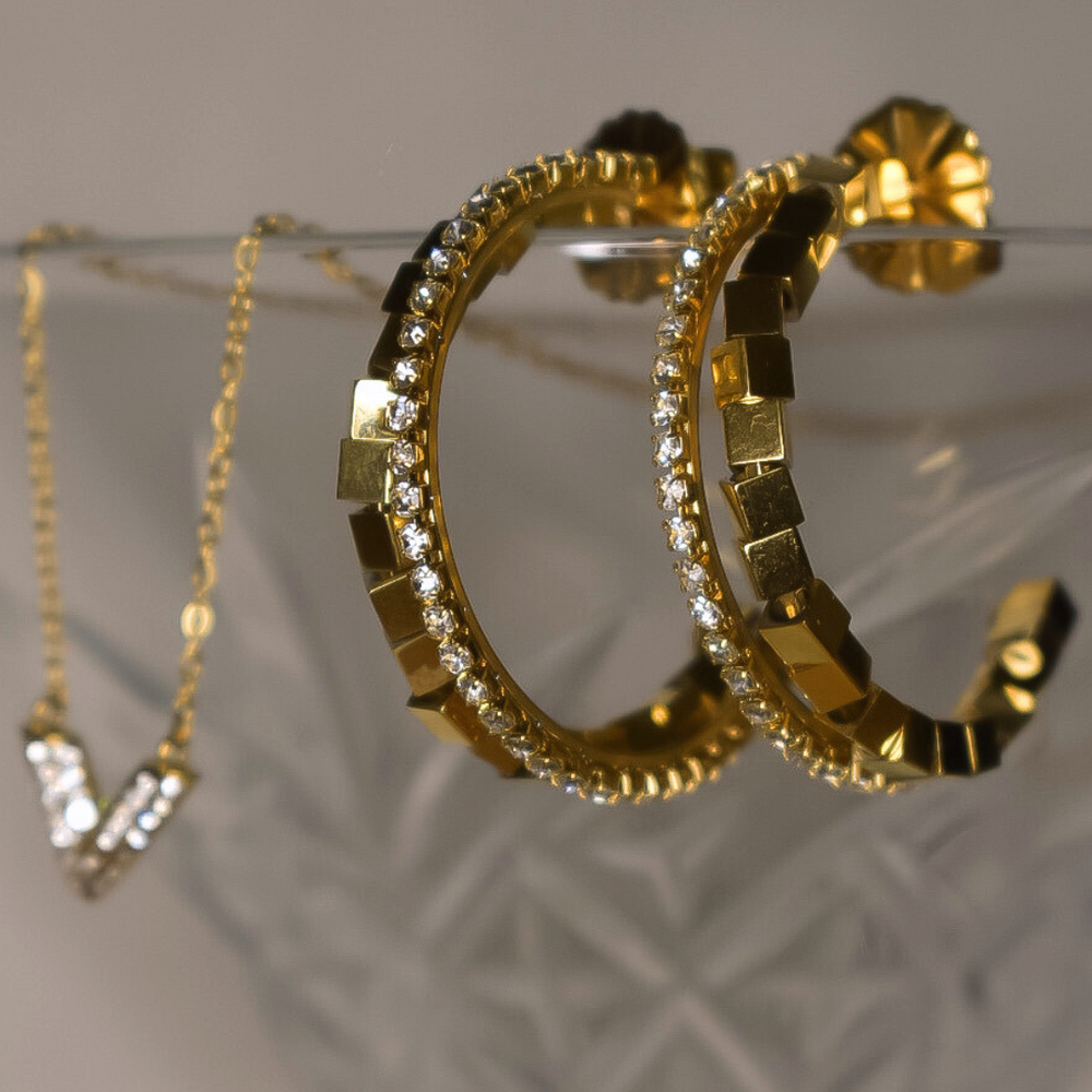 Close-up van de Lily oorbellen, met een gouden ringdesign en een rij van vierkante gouden kraaltjes en zilveren stras steentjes, gemaakt van stainless steel, hypoallergeen, waterproof en verkleuren niet.